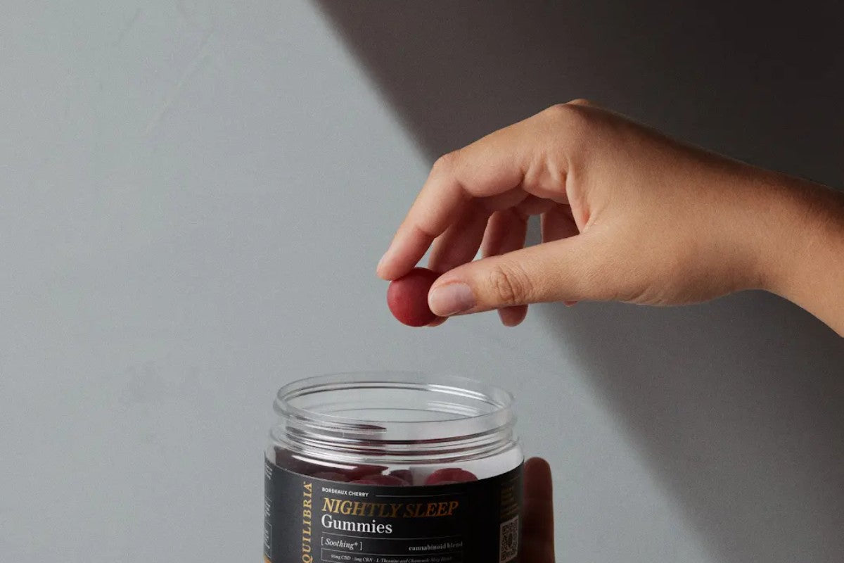 Hand holding a single red CBD softgel capsule above a clear glass jar on a gray background, illustrating CBN sleep support supplements