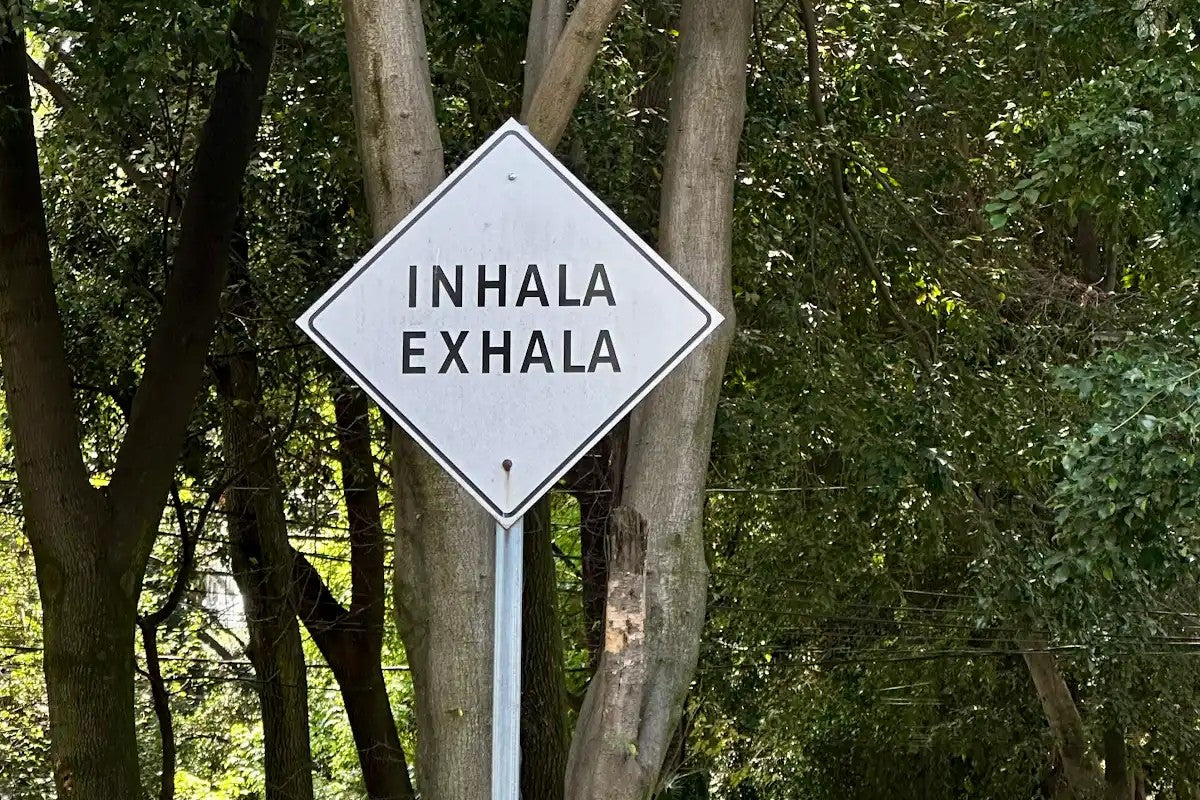 Outdoor diamond sign reading Inhala Exhala surrounded by lush green trees, symbolizing mindful breathwork and holistic wellness