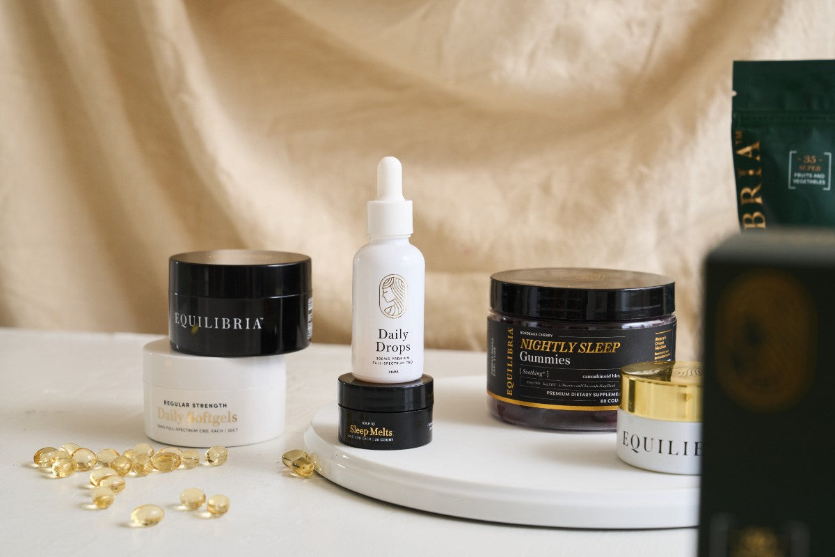 Equilibria Daily Drops CBD tincture centered in a flatlay of Equilibria wellness products including Daily Softgels and Nightly Sleep Gummies on a warm linen background