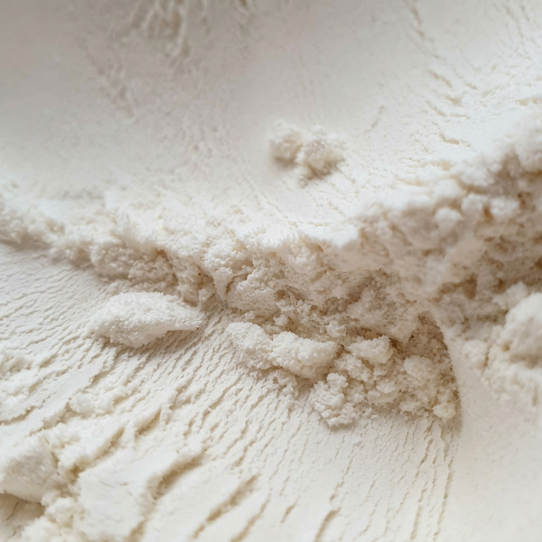 A close-up texture shot of an off-white powder, showing smooth ridges and soft clumps with a warm, slightly yellow undertone.