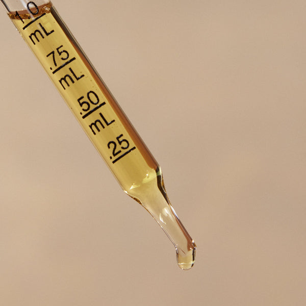 A close-up of a glass dropper filled with pale yellow liquid, angled diagonally against a solid beige background. The dropper has black measurement markings in milliliters and a single drop is hanging from the tip.