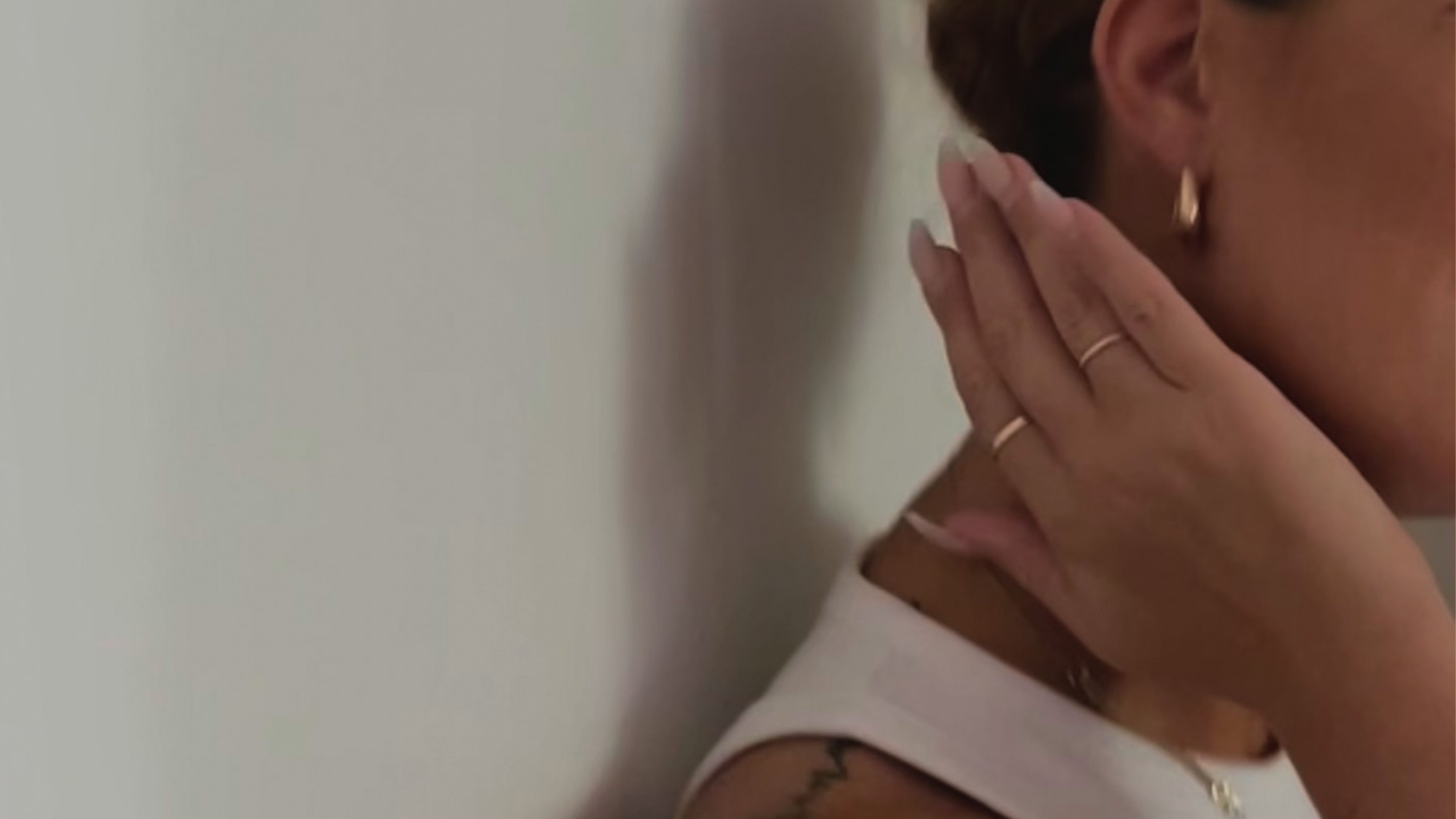 Close-up of a person wearing gold earrings and rings, holding their hand up to their neck, with a neutral background