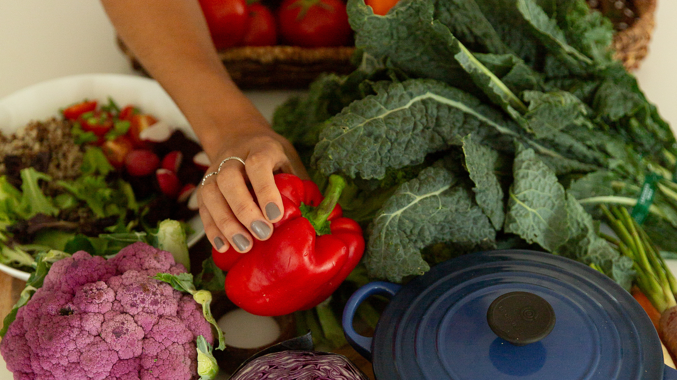 Image of a hand holding a red pepper above an abundant pile of various whole foods next to a dutch oven pot