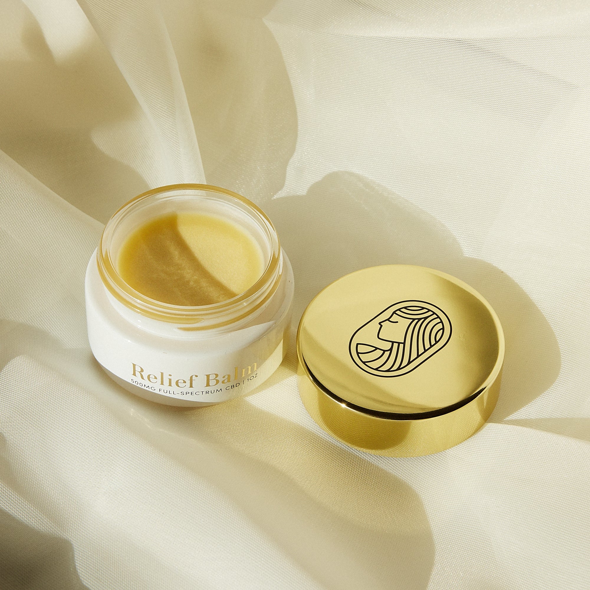 Jar of 'Relief Balm' with golden smooth balm and a gold lid on a soft beige background