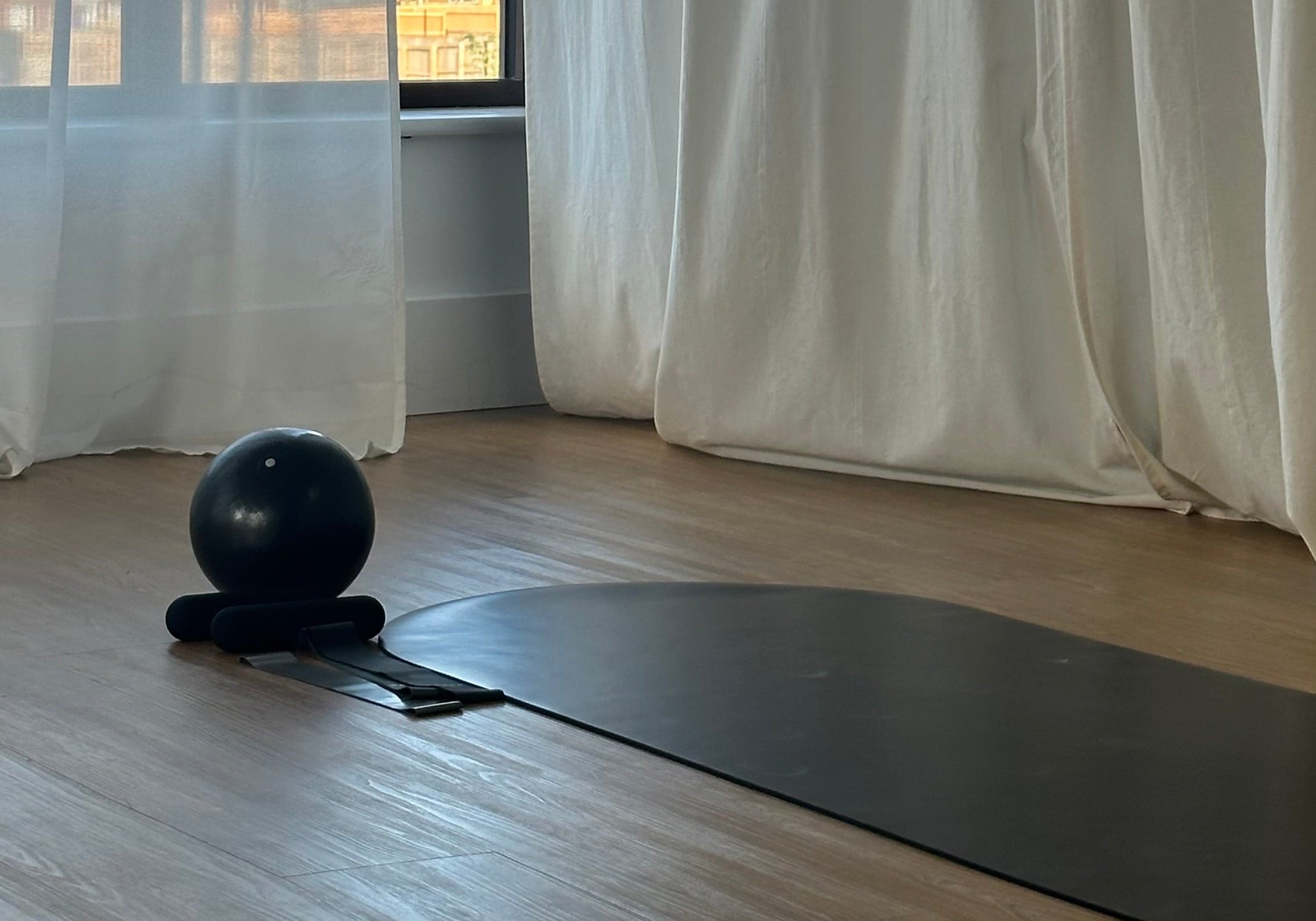 A black yoga mat and a black exercise ball on a wooden floor in a bright room with white curtains and a window.